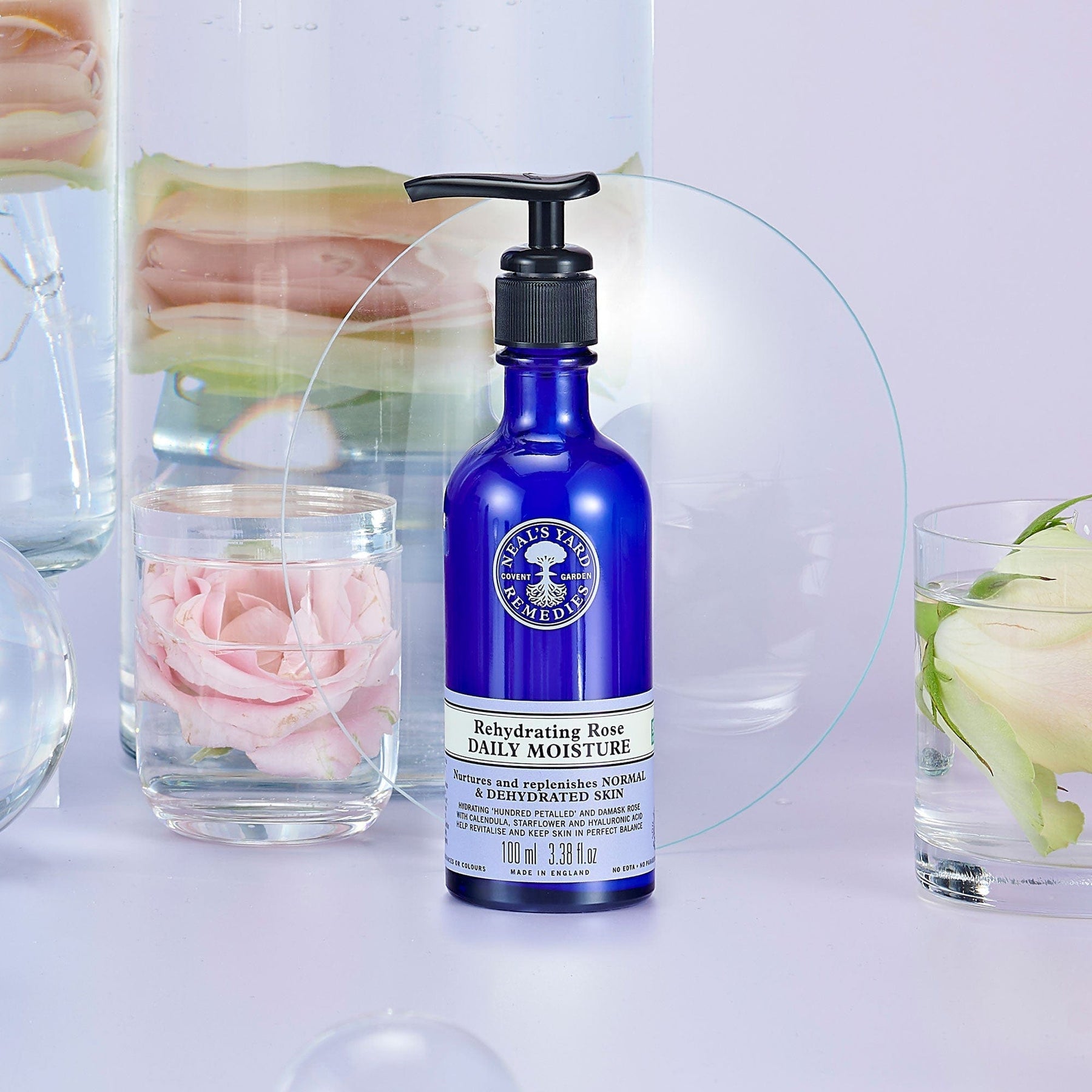 Neal's Yard Remedies Rehydrating Rose Daily Moisture in a blue bottle with beautiful purple background surrounded by flower petals in glasses.